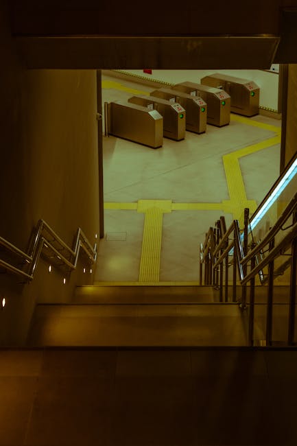 The image shows a downward view from a staircase inside a building leading to an underground or lower-level area at Bromley North Station, with visible tactile paving strips on the floor for accessibility. At the bottom of the stairs, there are four cardboard boxes with green and red indicators, positioned near automatic ticket barriers or access control gates, which are beige with metallic accents. The surrounding environment appears well-lit, with the area of movement clearly marked for safety during packing and home relocation tasks. The visual suggests a setting involving the logistical aspects of moving, such as coordinating transportation or loading items for a house removal service, with the branding of Removal Companies Bromley subtly implied by the context.