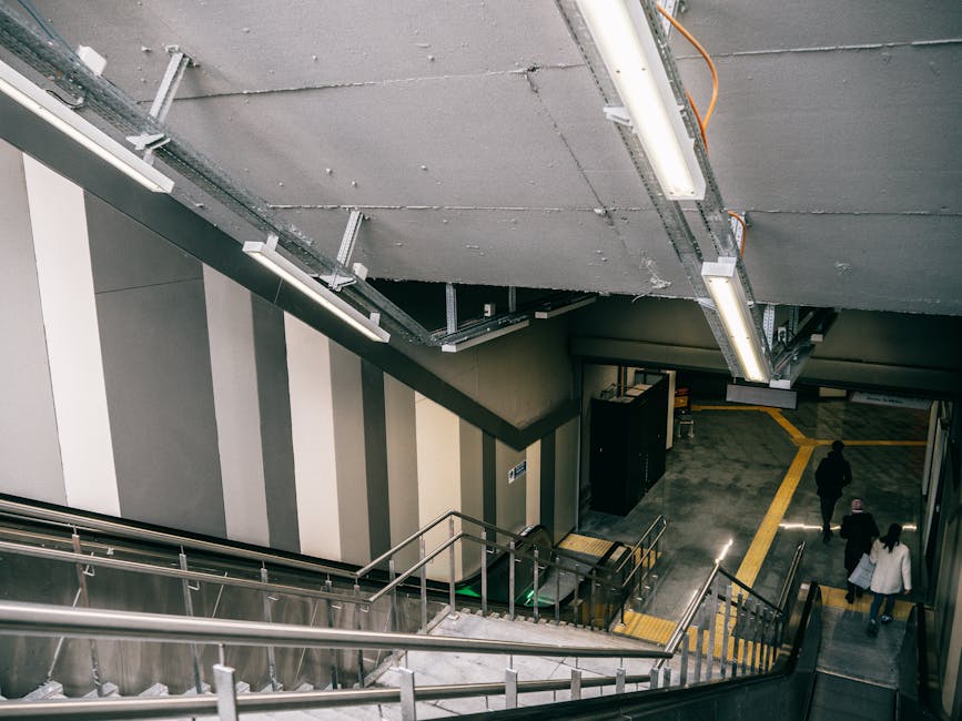 The image shows an indoor stairwell area at Bromley North Station used during a home relocation process. The staircase features metallic handrails and steps leading down to a tiled floor with yellow directional lines. Overhead, exposed ceiling panels and fluorescent lighting are visible, with some wiring and electrical conduits. In the background, three individuals are walking toward an exit, carrying bags and personal belongings. Adjacent to the stairs, there is a black cabinet or electronic equipment. The environment is well-lit, clean, and organized, and the overall scene depicts the loading or unloading phase in a train station setting, consistent with furniture transport or packing and moving activities associated with professional removal services provided by Removal Companies Bromley as part of their removals service category.