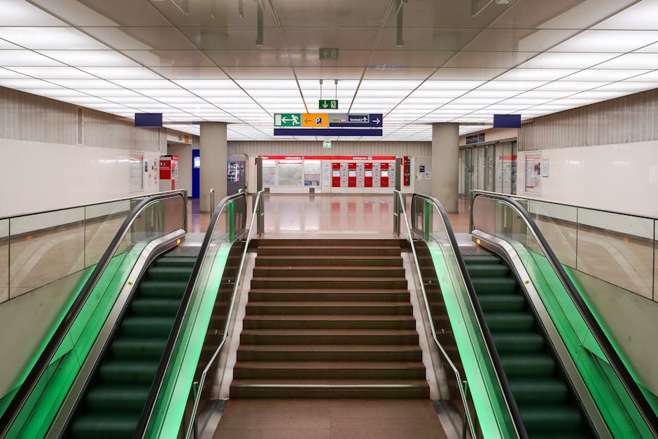 The image shows a downward view from a staircase inside a building leading to an underground or lower-level area at Bromley North Station, with visible tactile paving strips on the floor for accessibility. At the bottom of the stairs, there are four cardboard boxes with green and red indicators, positioned near automatic ticket barriers or access control gates, which are beige with metallic accents. The surrounding environment appears well-lit, with the area of movement clearly marked for safety during packing and home relocation tasks. The visual suggests a setting involving the logistical aspects of moving, such as coordinating transportation or loading items for a house removal service, with the branding of Removal Companies Bromley subtly implied by the context.
