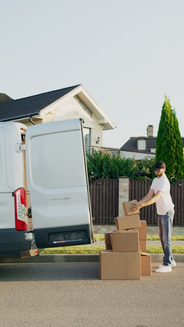A young man wearing casual clothing with a beard is standing outside a residential property during daytime, arranging cardboard boxes for a home relocation. He is stacking and preparing the boxes, which appear to be filled with household items, next to a white van with its rear doors open, positioned on the driveway. The van is used for furniture transport and packing and moving purposes, with the open cargo area revealing additional boxes inside. Behind the house, there is a fence, well-maintained garden, and tall green trees, all under a clear sky with natural daylight illuminating the scene. The image captures the loading process, with the man handling the boxes to facilitate efficient removal and moving services, consistent with the professional offerings of Removal Companies Bromley and their affordable, honest pricing approach as described on the webpage 'Cheap Bromley removals BR1 BR2 honest pricing explained'.