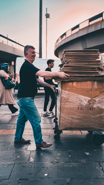 A man wearing a black shirt and blue jeans is pushing a large, upright trolley loaded with flattened cardboard boxes and wrapped packaging materials, likely in preparation for a house removal or moving process. He is outside under a bridge or elevated roadway, with other pedestrians visible nearby, some walking in the background. The trolley is connected to a collection of packed boxes and household items, indicating an organized packing and moving operation. The environment is urban with paved ground and a tall pole with attached equipment. The scene reflects the practical aspects of furniture transfer and home relocation handling, with Removal Companies Bromley providing professional removals services. The overall setting captures the logistics involved in loading and transporting belongings for a home move, with visible packaging materials and equipment supporting efficient packing and transfer tasks.