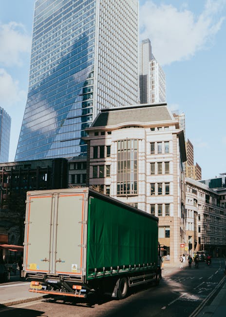 A large delivery truck with a green cargo area is parked on a city street adjacent to tall modern glass skyscrapers and older stone buildings. The truck is positioned at the curb, with its rear doors closed, and appears to be in the process of loading or unloading. The surroundings include a pavement with pedestrians and cyclists, commercial signage, and urban infrastructure. The scene is captured in bright daylight with clear skies, emphasizing the urban environment where professional removal and moving services provided by companies like Removal Companies Bromley might operate during a home relocation or furniture transport process.
