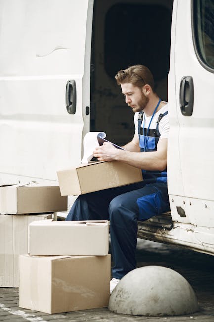 A male removal worker with a beard, dressed in a striped shirt and blue overalls, is sitting on the edge of a white moving van with its side door open. He is holding a cardboard box on his lap while reviewing a clipboard or list, preparing for home relocation. Surrounding him on the ground are several other cardboard boxes of various sizes, some stacked and others placed on the pavement. The scene is set outdoors near a curb, with a concrete bollard visible in the foreground, indicating an urban environment. Inside the van, visible behind the worker, are more packing materials and possibly additional boxes or furniture being loaded or unloaded as part of the furniture transport and packing process. The lighting appears natural, suggesting daytime, consistent with professional removals and moving logistics activities carried out by companies like Removal Companies Bromley.